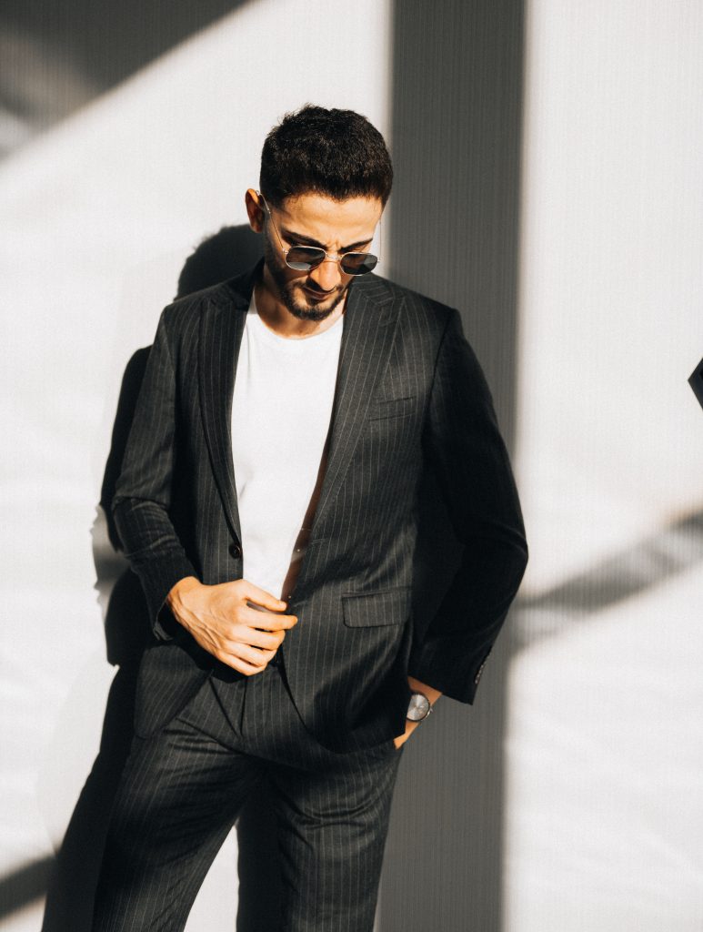 A man in a pinstriped suit, white t-shirt, and sunglasses posing against a sunlit wall.