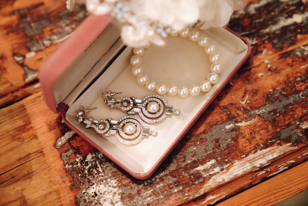 A high-angle close-up of a rustic wooden surface with a pink jewelry box displaying a pair of ornate silver drop earrings.