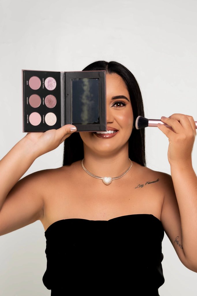 Woman holding a makeup palette and brush wearing a black strapless dress