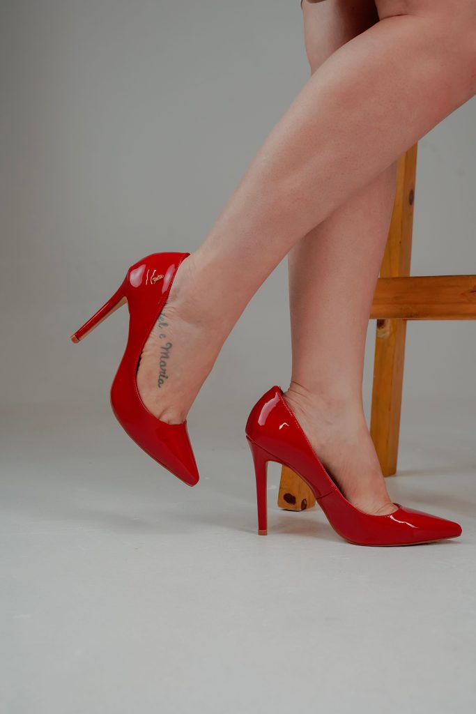 A close-up focus on a person's legs wearing bright red pointed-toe high heels against a minimalist backdrop.
