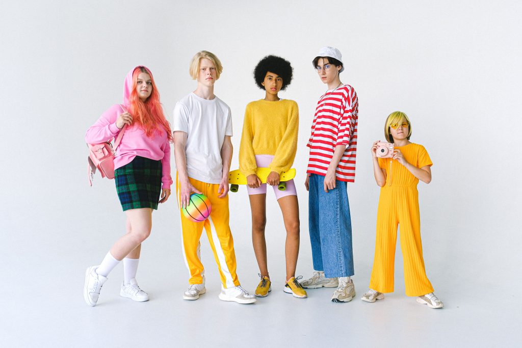 Five young people posing together against a white backdrop in bright, colorful streetwear including yellow trousers, striped tops, and pink apparel.