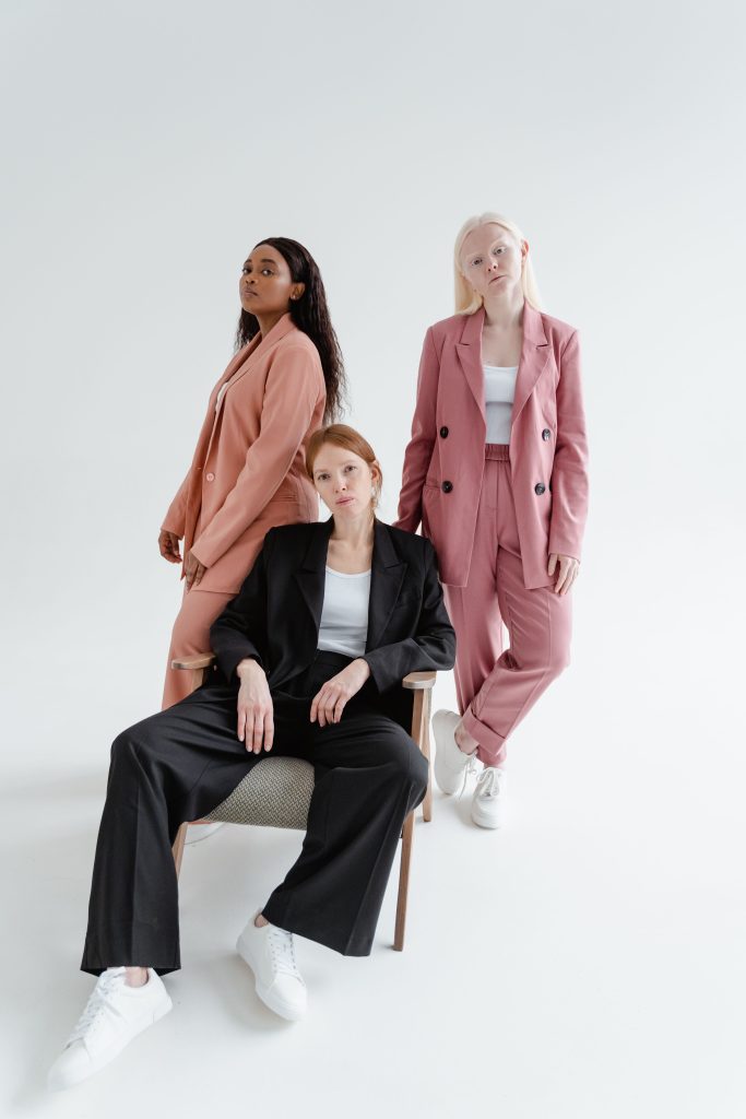 Three women modeling tailored pant suits against a white backdrop—one seated in front wearing a charcoal grey suit, and two standing behind her wearing pink suits.